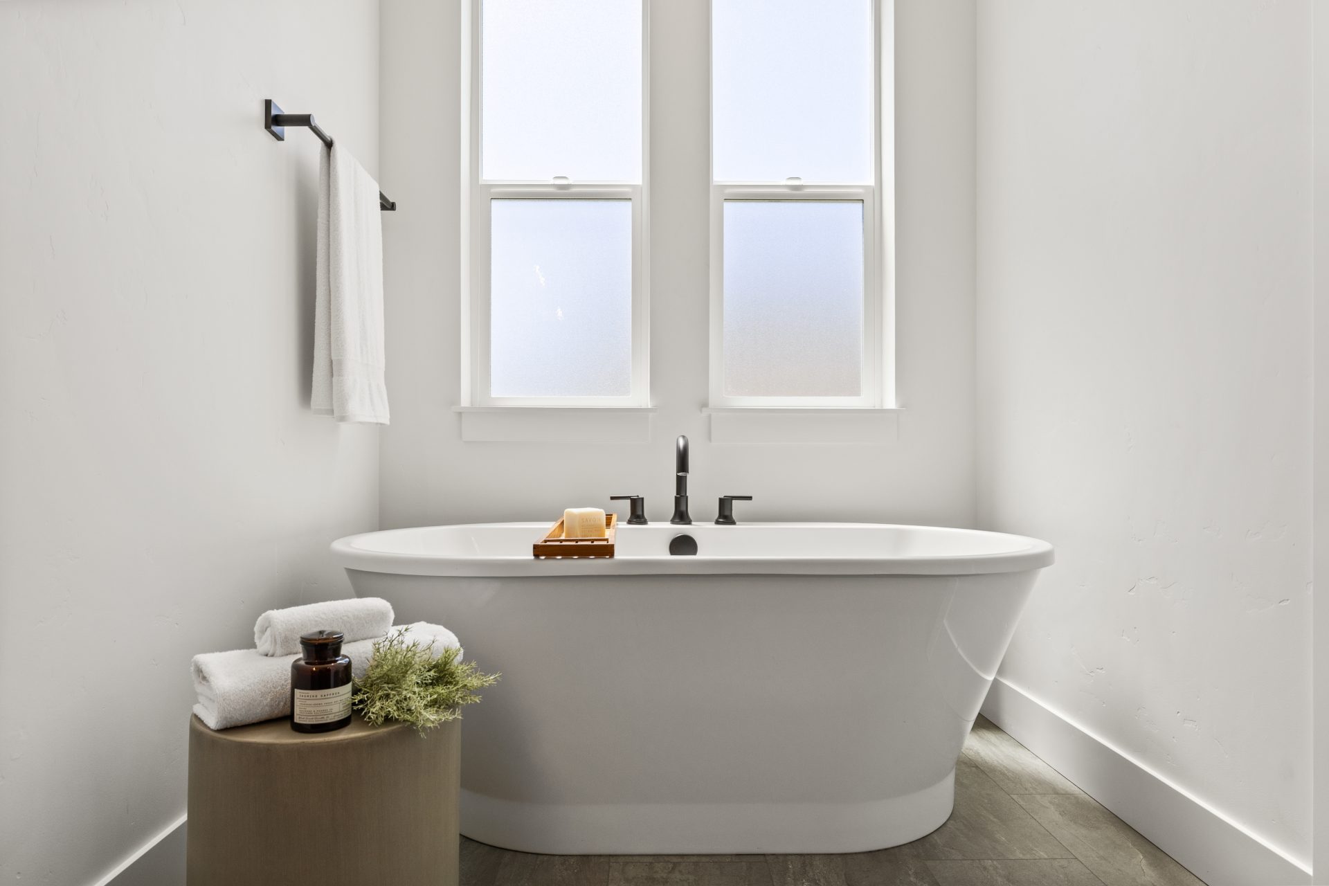 Bathroom with a freestanding soaking tub and a contemporary design. The space features a black faucet and towel bar, a frosted-glass window, and a wood side table with rolled towels and decorative accents against white walls.