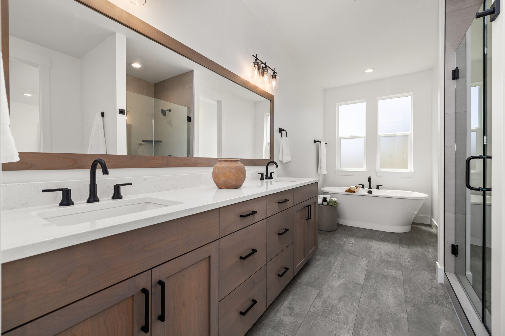 Primary bathroom with tile flooring and a contemporary design. The space features a wood double-vanity with white countertops, black faucets, and a large wood-framed mirror. A freestanding soaking tub sits beneath a window, with a glass-enclosed shower to