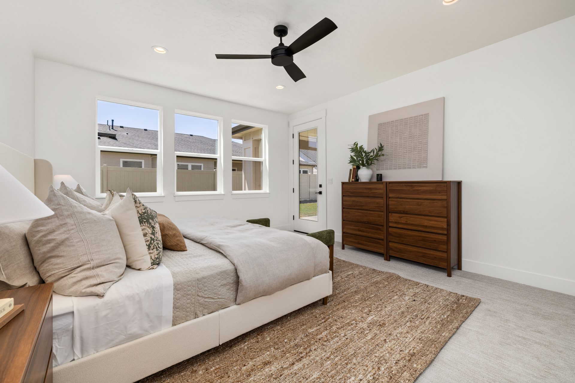 Primary bedroom with carpeted flooring and a contemporary design. The room features an upholstered bed with accent pillows, a wood dresser with framed wall art, a woven area rug, and a black ceiling fan overhead. Large windows and a door with glass panels