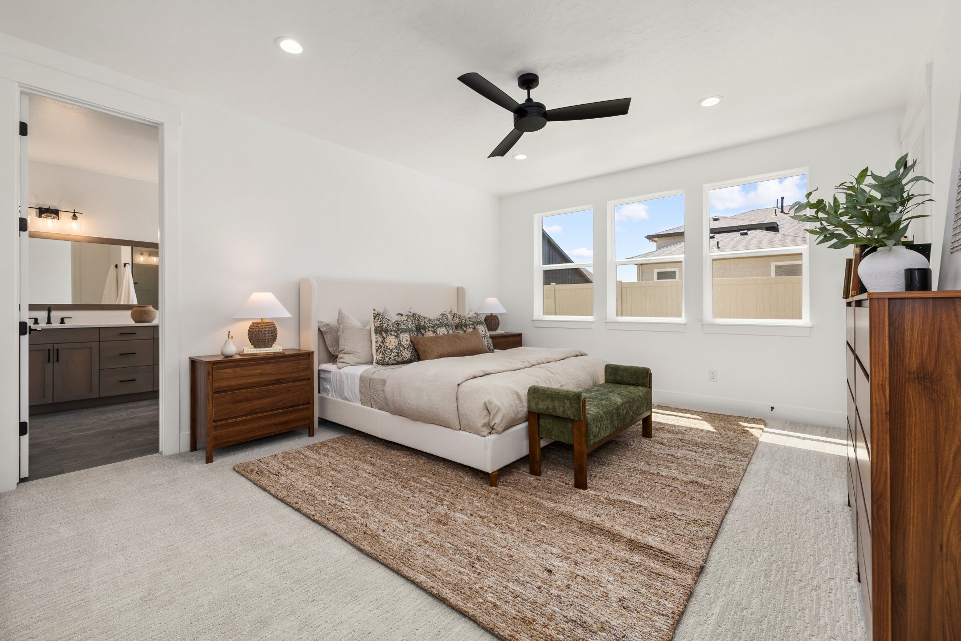 Primary bedroom with carpeted flooring and a contemporary design. The room features an upholstered bed with accent pillows, wood nightstands with lamps, a woven area rug, and a black ceiling fan overhead. A bench sits at the foot of the bed, large windows