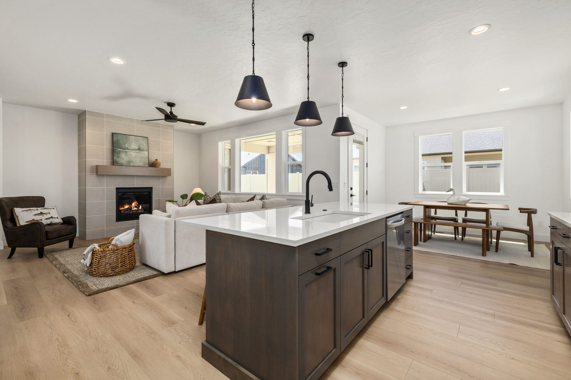 Open-concept main living area with light hardwood flooring and a contemporary design. The kitchen features a large center island with a dark wood base, white countertop, built-in dishwasher, and black pendant lighting. The space flows into a living room w