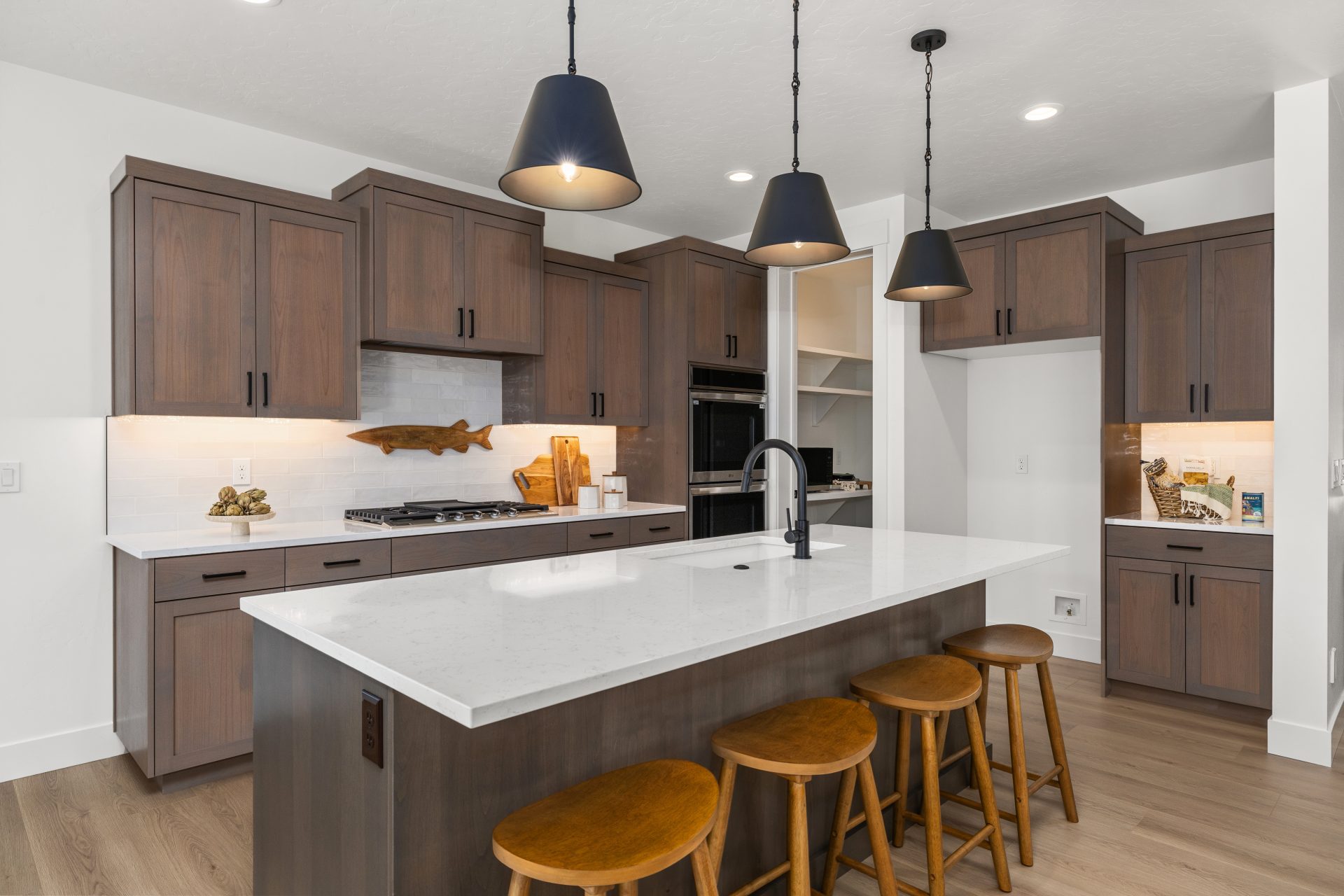 Kitchen with light hardwood flooring and a contemporary design. The space features wood cabinetry, a large center island with a white waterfall countertop, three wood bar stools, and black pendant lighting. A built-in oven, gas cooktop, and tile backsplas