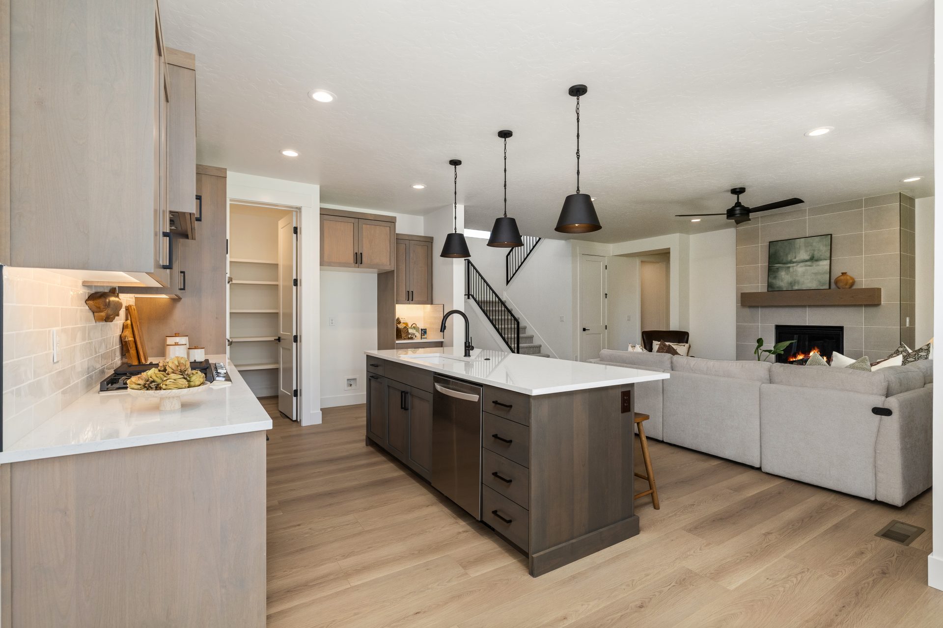 Kitchen with light hardwood flooring and a contemporary design. The space features two-tone cabinetry, a large center island with a dark wood base and white countertop, a built-in dishwasher, and black pendant lighting. The kitchen opens to a living area 