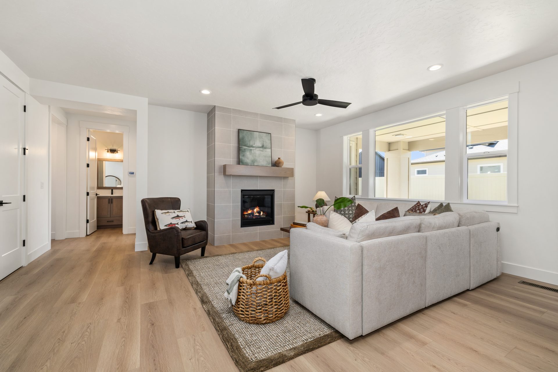Living room with light hardwood flooring and a contemporary design. The space features a tile-surround fireplace, a sectional sofa with accent pillows, a leather accent chair, and a woven area rug, with a black ceiling fan overhead. A hallway leads toward