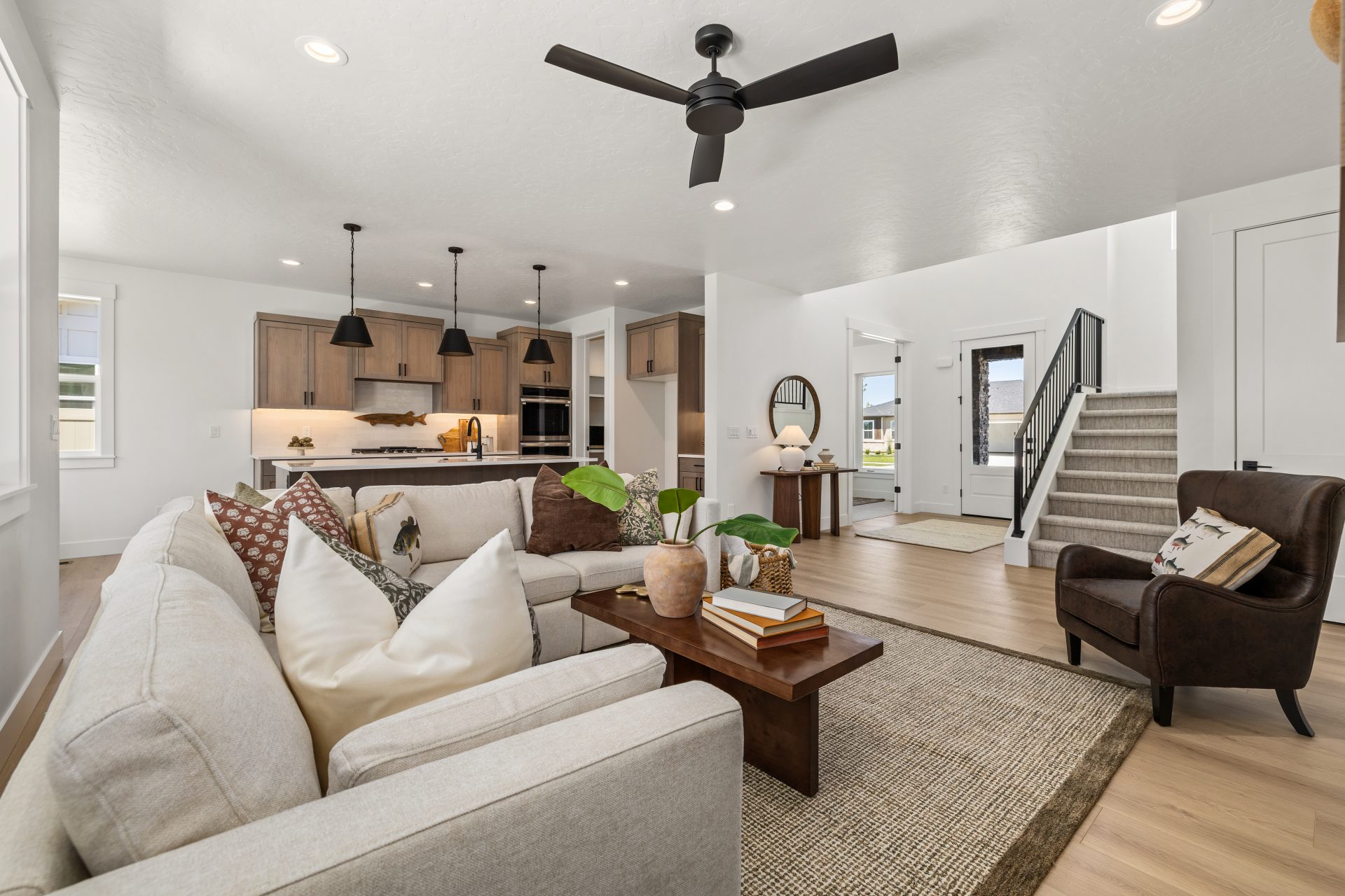 Open-concept living space with light hardwood flooring and a contemporary design. The living room features a sectional sofa with accent pillows, a leather accent chair, a wood coffee table, and a woven area rug, with a black ceiling fan overhead. The spac
