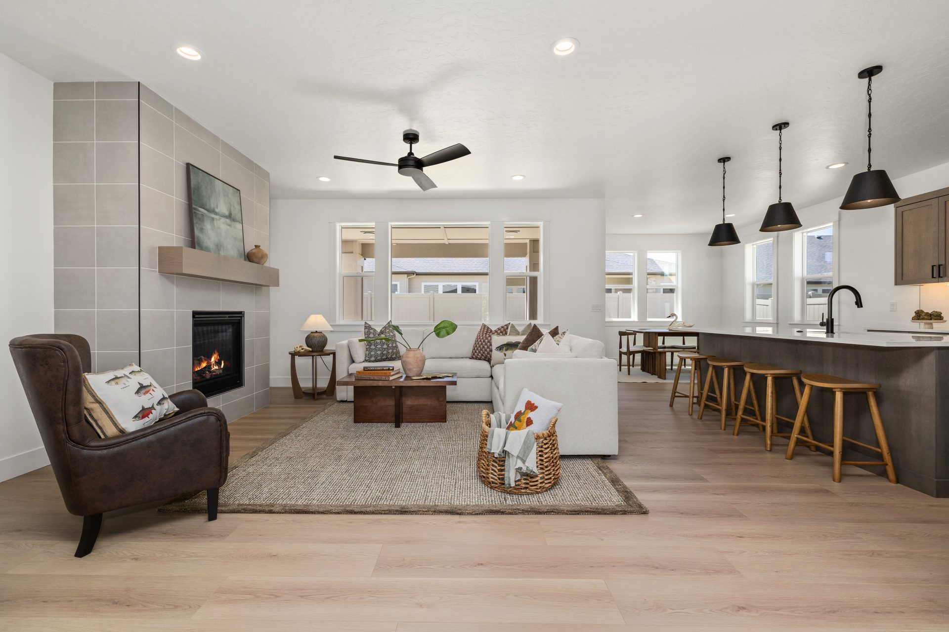 Open-concept living space with light hardwood flooring and a contemporary design. The living room features a tile-surround fireplace, a sectional sofa, a leather accent chair, and a woven area rug, with a ceiling fan overhead. The space flows into a kitch