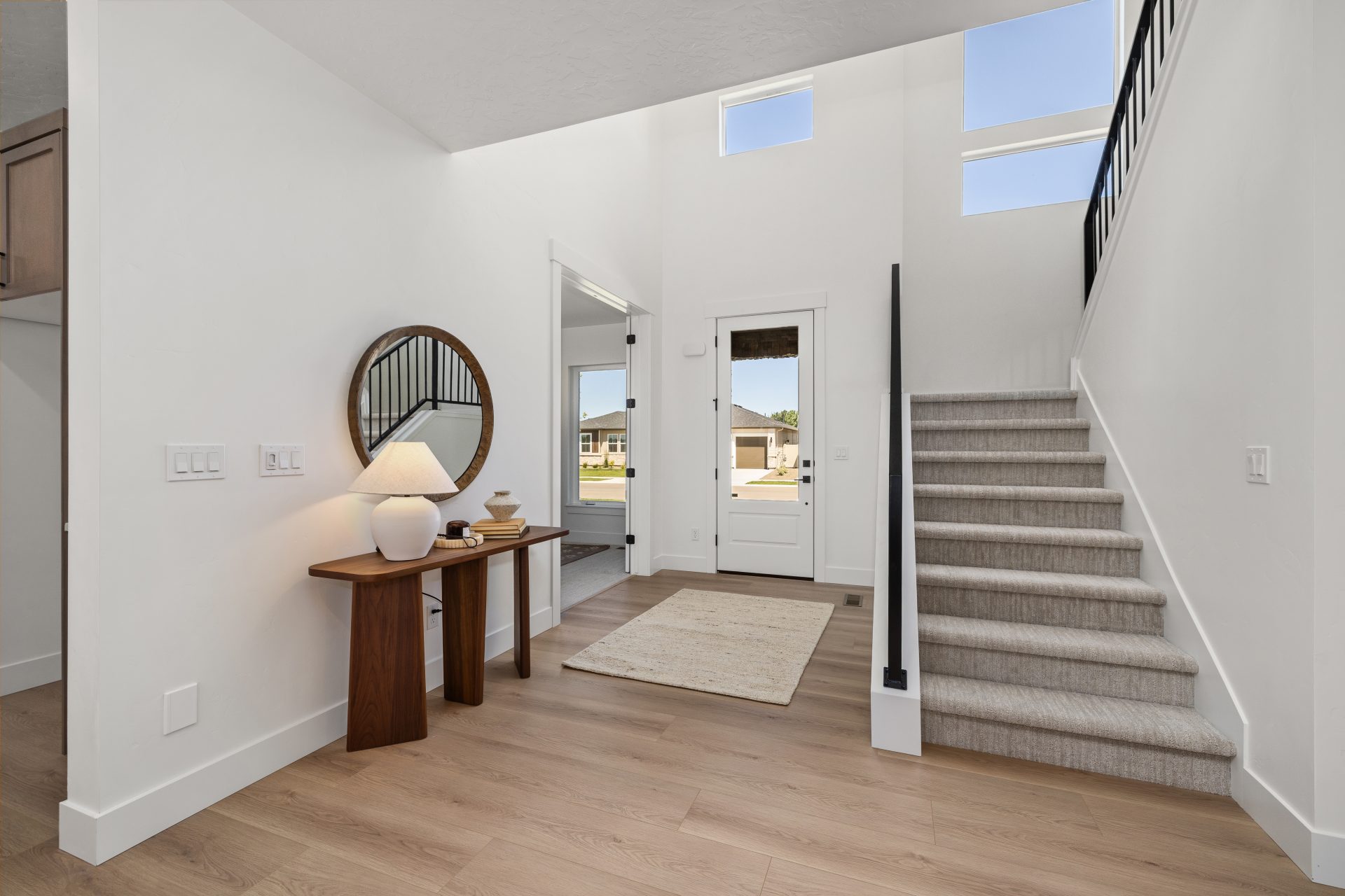 Two-story home entryway with a high ceiling and abundant natural light from upper-level windows. The space features light hardwood flooring, a wood console table with a round mirror and lamp, and a neutral runner rug. A carpeted staircase with a black met