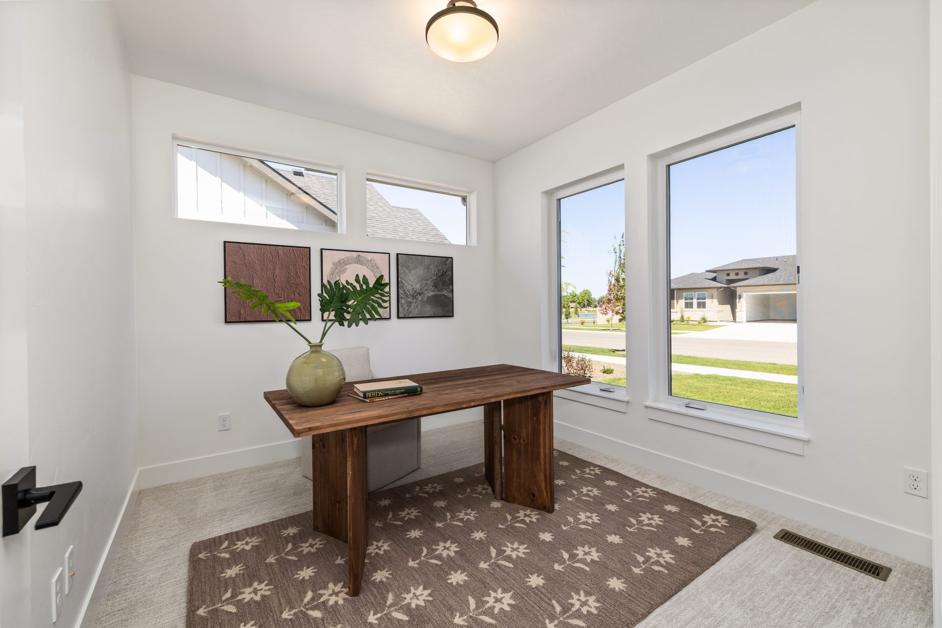 Home office with a wood desk and green vase, framed wall art, a patterned area rug, and large windows letting in natural light.
