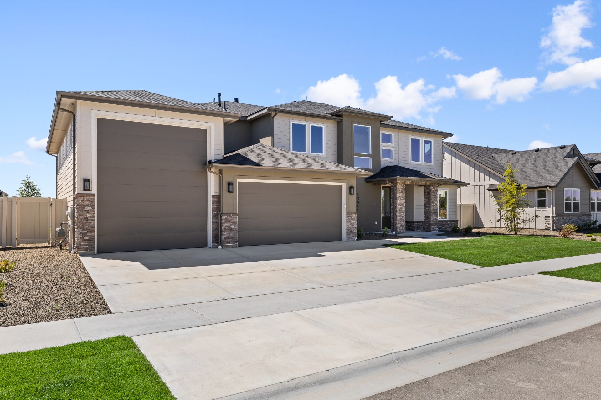 Two-story suburban home with taupe siding and stone accents, a three-car garage, a wide paved driveway, and a landscaped front lawn under a clear blue sky.