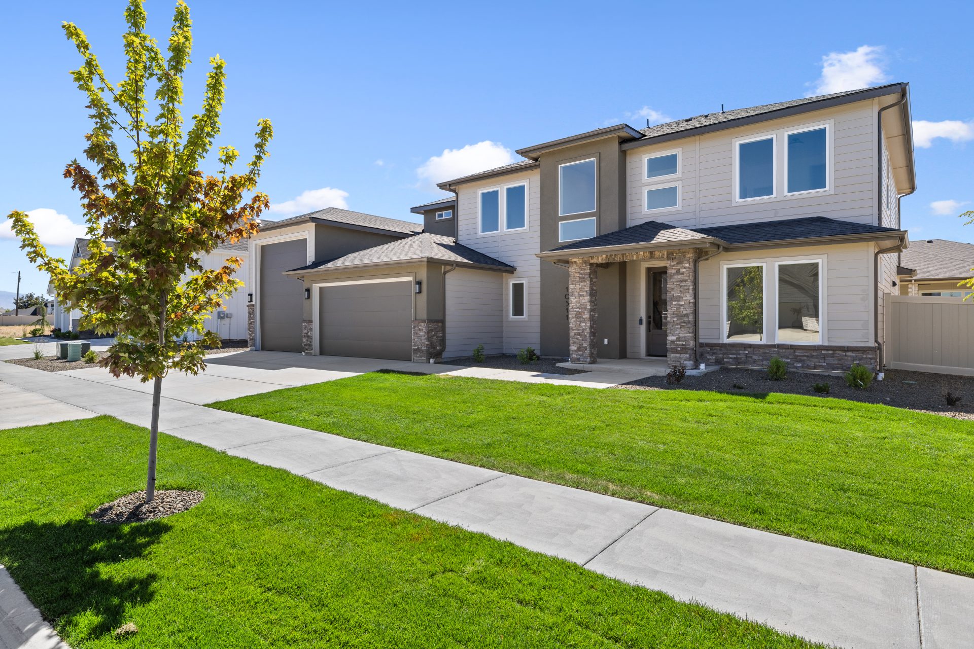 Two-story suburban home with tan and gray siding, a stone-accented entryway, an attached two-car garage, a paved driveway, and a green front lawn with a young maple tree under a clear blue sky.