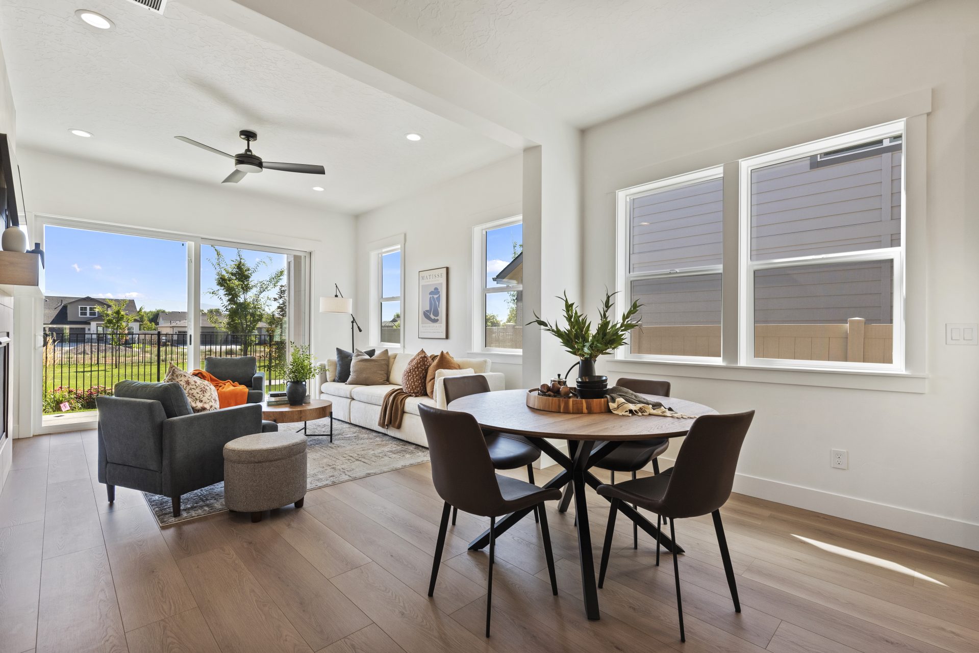 Open-concept living and dining area with light hardwood flooring and a contemporary design. The space features a round dining table with black chairs, a sofa and accent chairs in the living area, and a black ceiling fan overhead. Large windows and a slidi