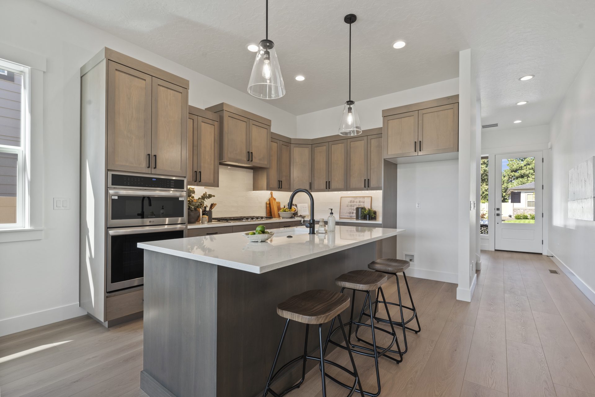 Kitchen with light hardwood flooring and a contemporary design. The space features wood cabinetry, a large center island with a dark wood base and white countertop, bar stools, and glass pendant lighting. A built-in oven, gas cooktop, and tile backsplash 